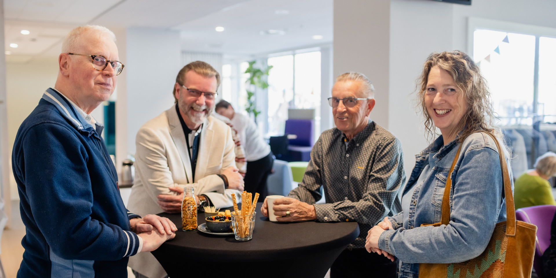 Groep mensen (huurdersvereniging en collega's Staedion) lachen en praten rond een statafel in een binnenruimte.