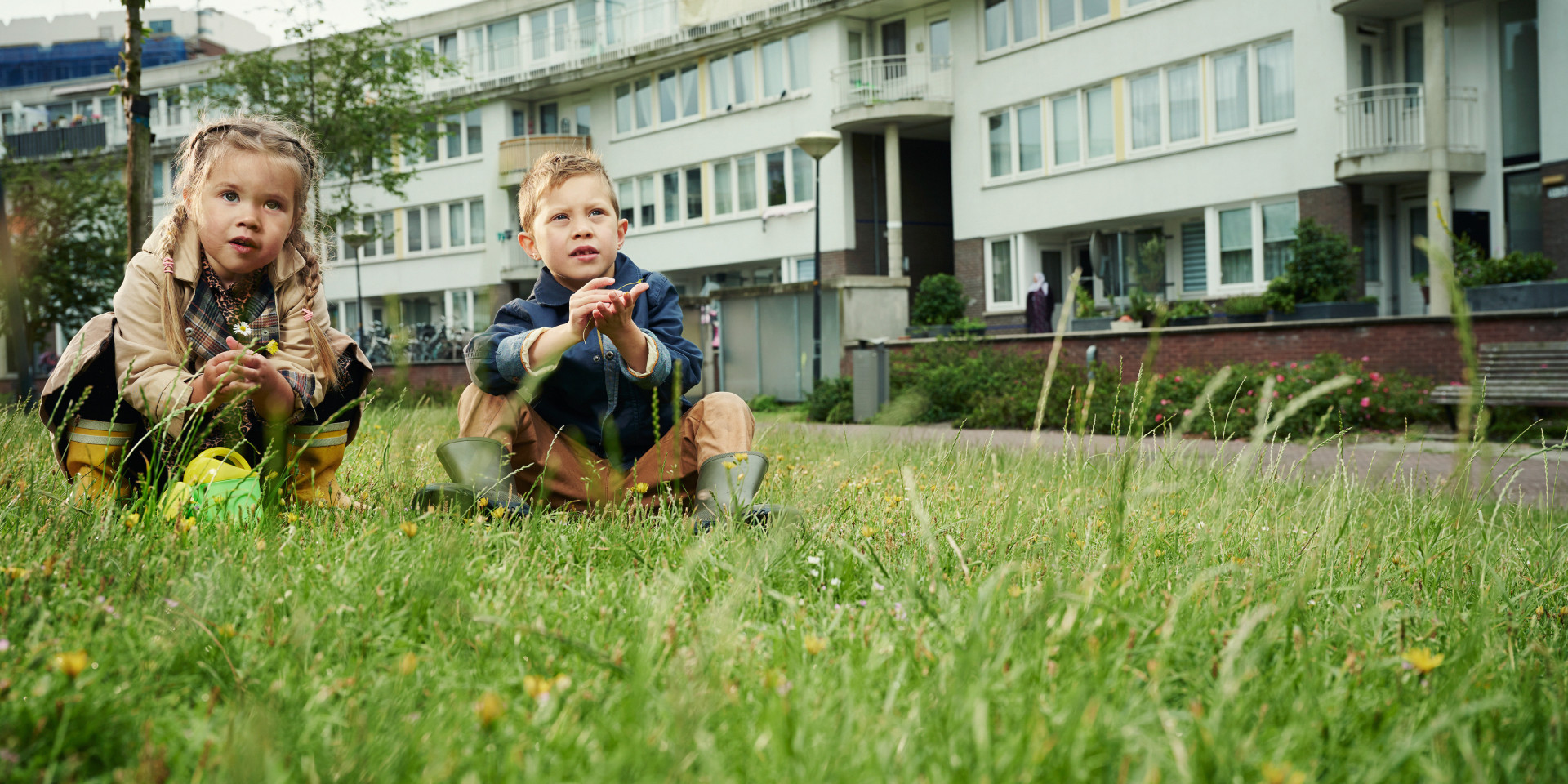 Twee kinderen zittend in het gras voor een rij flatgebouwen.