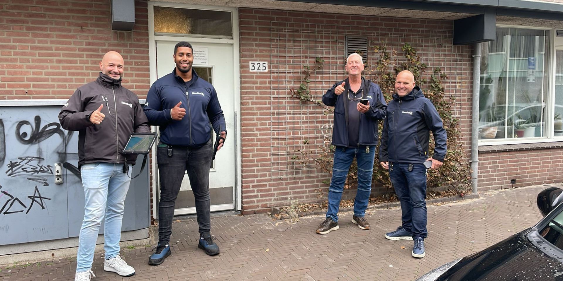 Groep collega's van Staedion poseren voor een gebouw, glimlachend en duim omhoog stekend.