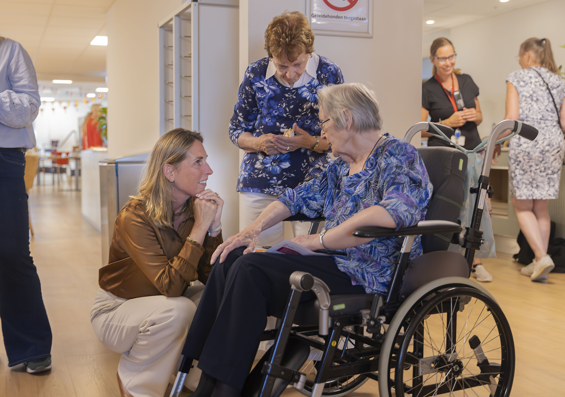 Marieke Stam knielt en praat met oudere dame in rolstoel.